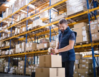 Male warehouse worker sealing cardboard boxes with racking in background